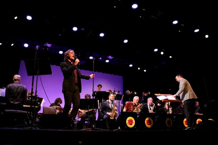 A live performance featuring a singer in a suit holding a microphone, accompanied by a jazz band with brass instruments and a pianist. A conductor stands to the right, leading the ensemble on a stage illuminated by bright overhead lights.